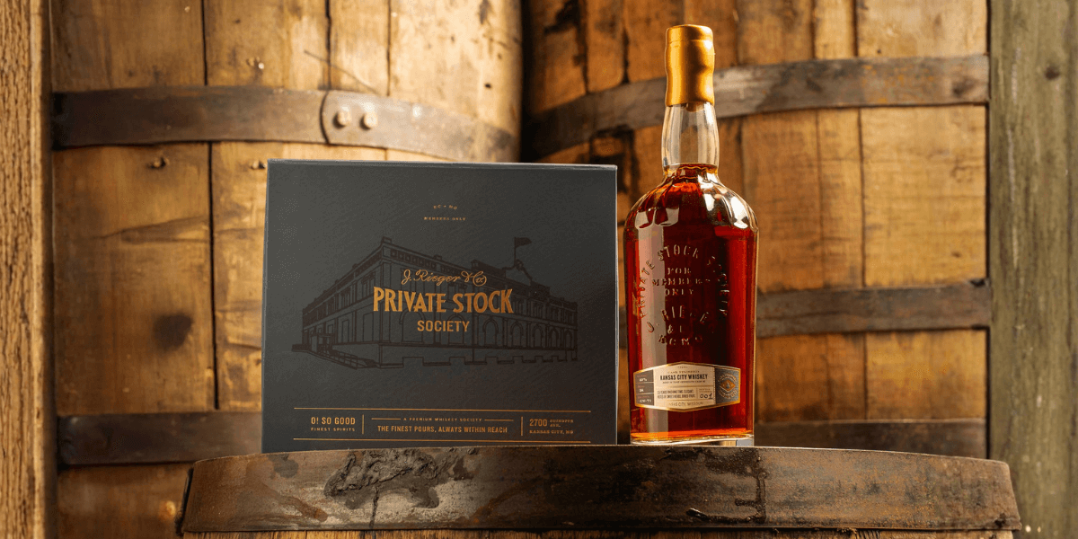 J. Rieger & Co. Private Stock Kansas City Whiskey bottle and premium gift box displayed on a whiskey barrel with rustic wooden backdrop.