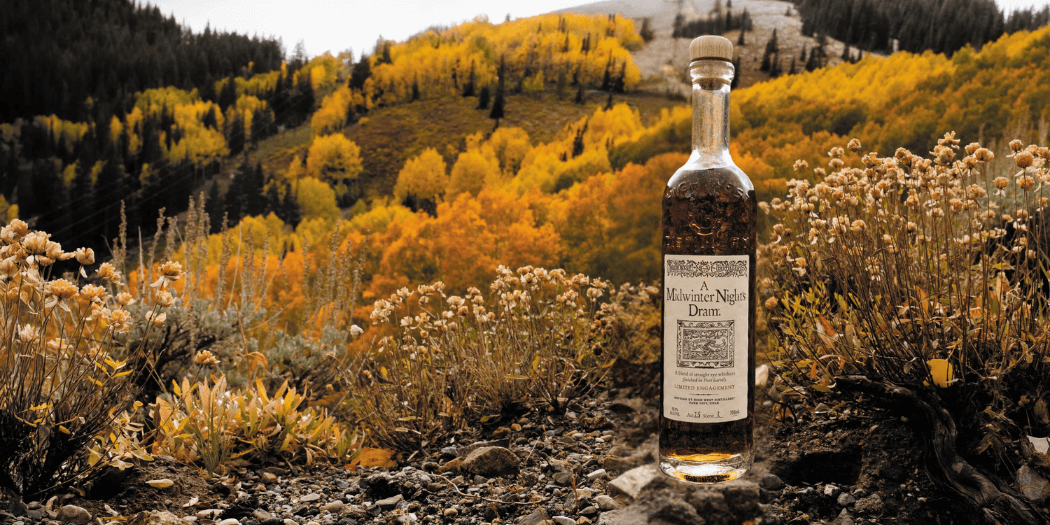 Bottle of A Midwinter Night’s Dram® Act 13 standing on rocky terrain with autumn foliage and a mountain in the background, capturing the seasonal essence of High West’s limited-release rye whiskey.