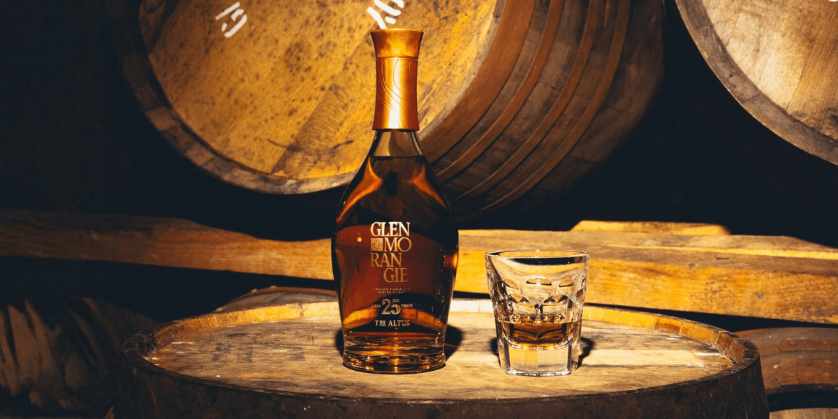 A bottle of Glenmorangie The Altus 25 Years Old Highland Single Malt Scotch Whisky and a filled glass sit on a wooden cask in a dimly lit barrel room, surrounded by aging oak barrels.