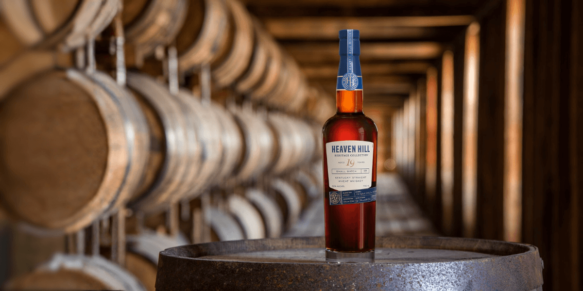 A bottle of Heaven Hill Heritage Collection 19-Year Kentucky Straight Wheat Whiskey displayed on a barrel inside a dimly lit rickhouse, with rows of aging barrels in the background.