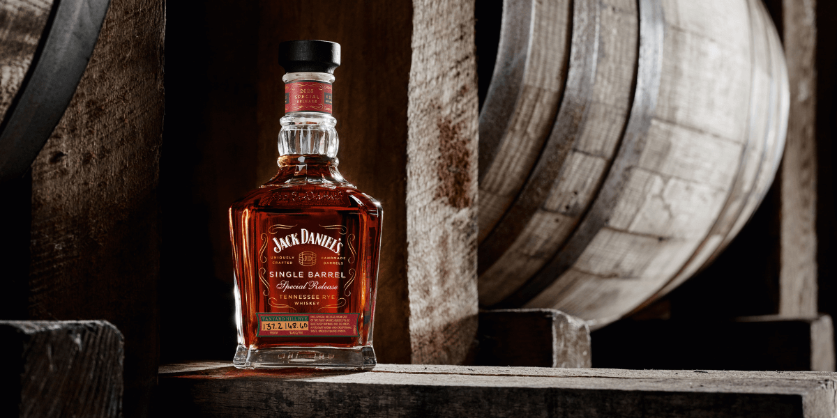 Jack Daniel’s Single Barrel Special Release 2025 Tennessee Rye Whiskey bottle displayed in a rustic barrelhouse, positioned beside aging oak barrels with warm, moody lighting highlighting the deep amber color of the whiskey.