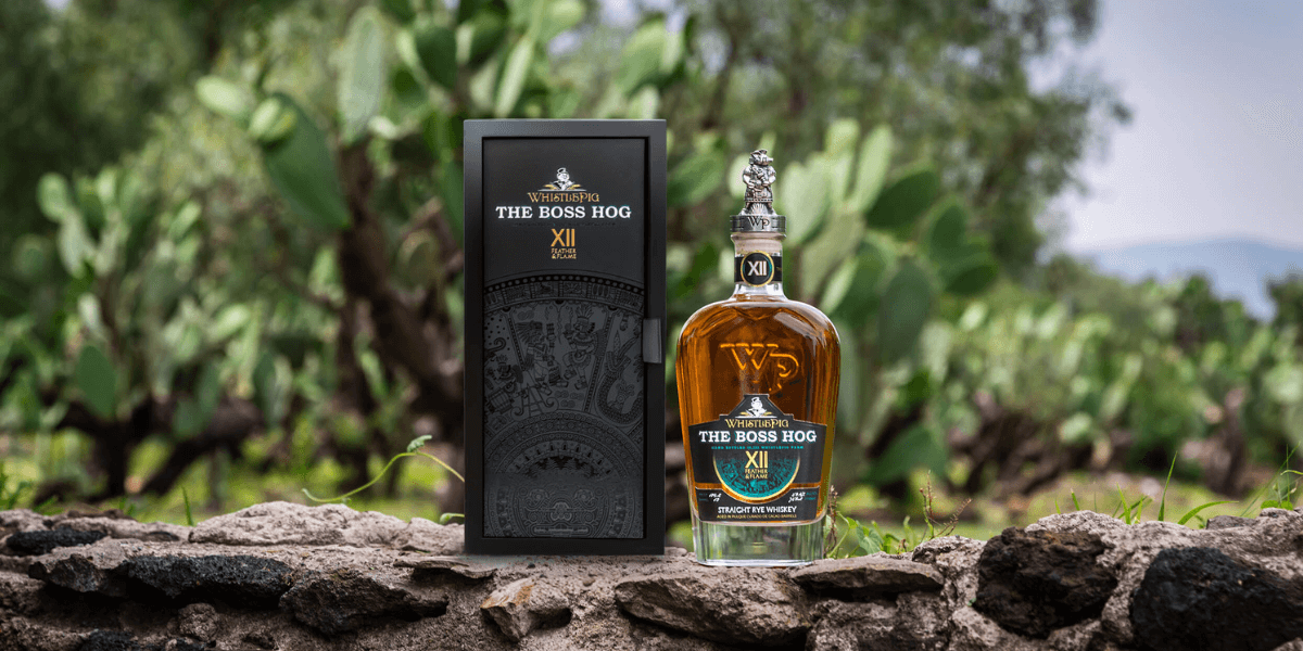 A bottle of WhistlePig The Boss Hog XII: Feather & Flame straight rye whiskey stands beside its black presentation box on a stone wall, set against a backdrop of green agave plants and an open sky.