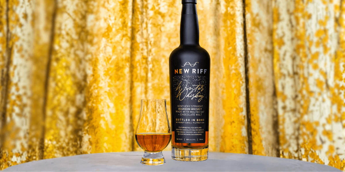 A bottle of New Riff Winter Whiskey and a Glencairn glass filled with bourbon, set against a gold velvet backdrop. The black label highlights “Winter Whiskey” and notes the use of malted oats and chocolate malt.