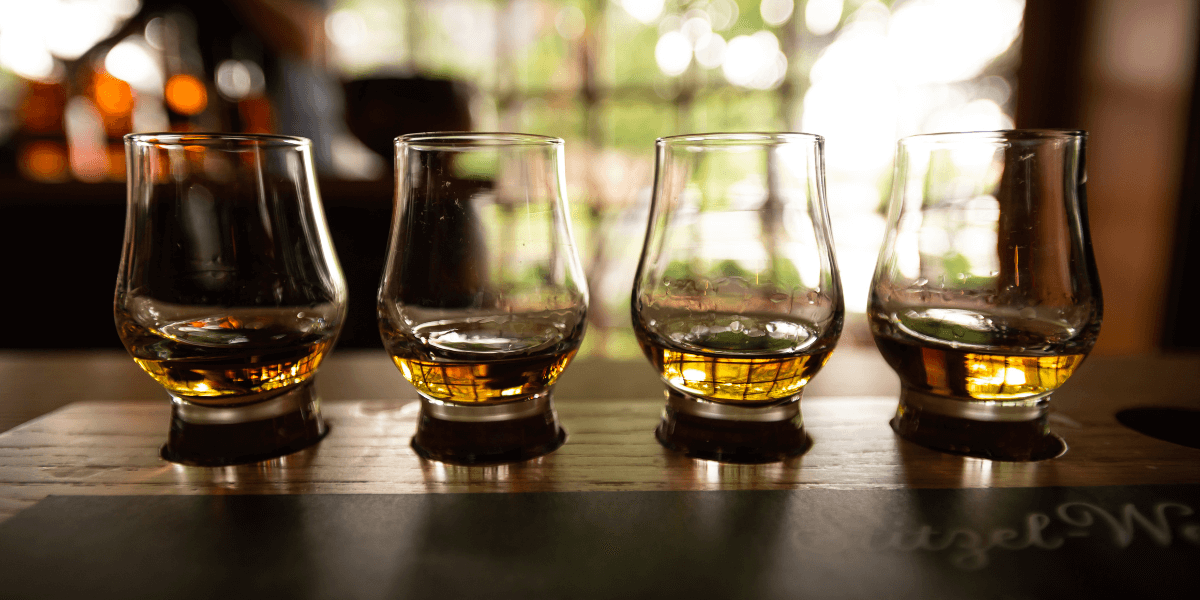 Four whiskey tasting glasses lined up on a wooden board with amber whiskey inside, prepared for a professional whiskey tasting.