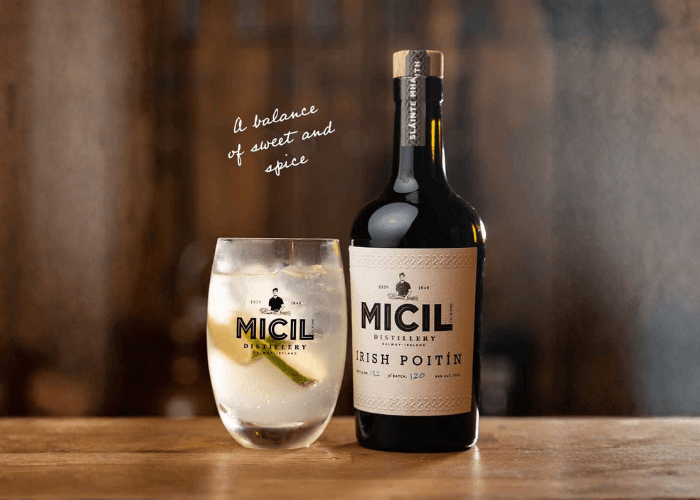 Micil Irish Poitín bottle alongside a served cocktail with ice and citrus in a branded glass on a wooden bar top