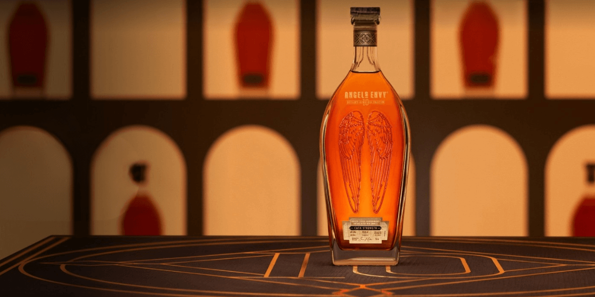 A bottle of Angel’s Envy 10 Cask Strength bourbon displayed upright on a patterned surface, with a warm, backlit background featuring arched cutouts and silhouettes of other bottles. The bottle’s signature etched angel wings are prominently visible through the amber whiskey.