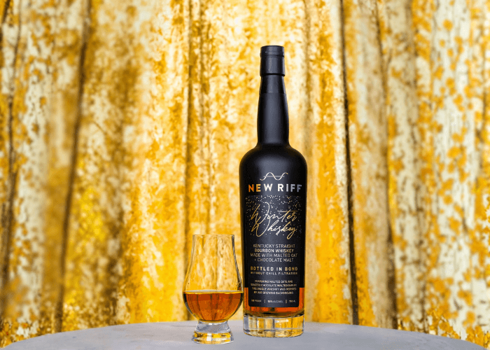 A bottle of New Riff Winter Whiskey and a Glencairn glass filled with bourbon, set against a gold velvet backdrop. The black label highlights “Winter Whiskey” and notes the use of malted oats and chocolate malt.