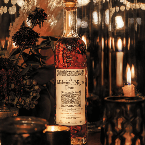Bottle of High West’s A Midwinter Night’s Dram® Act 13 surrounded by warm candlelight, dried flowers, and dark glassware, evoking a cozy, theatrical winter setting.