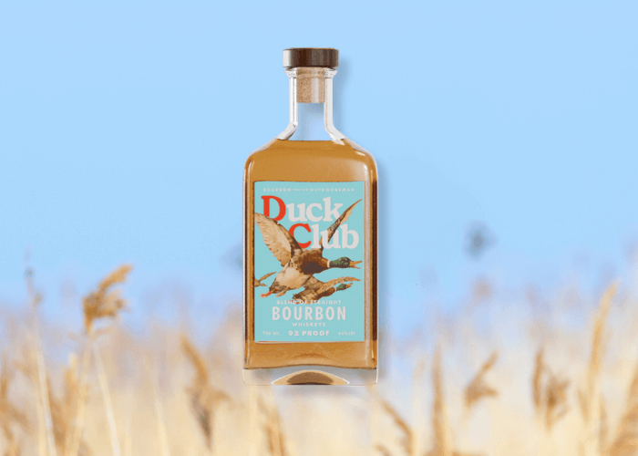 Bottle of Duck Club™ Bourbon “High Brass” 110 Proof standing in front of a blue sky with blurred golden reeds in the foreground.