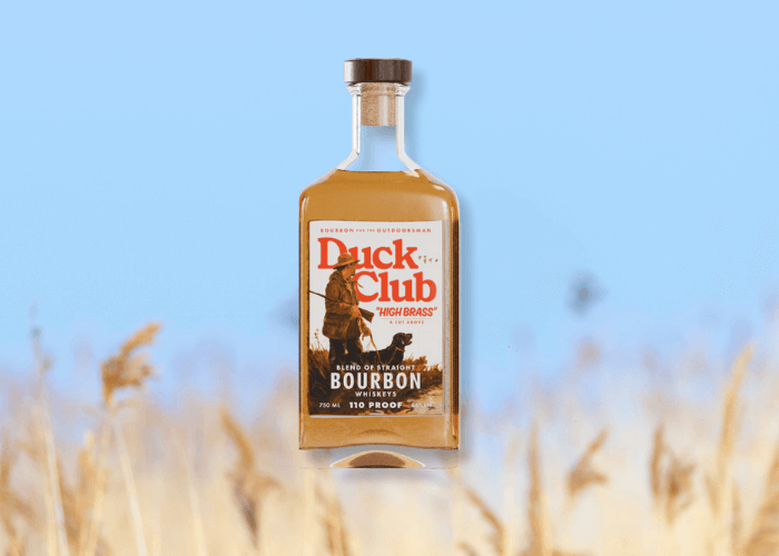 Bottle of Duck Club™ Bourbon 92 Proof featuring flying ducks on the label, set against a soft blue sky with blurred wheat in the foreground.