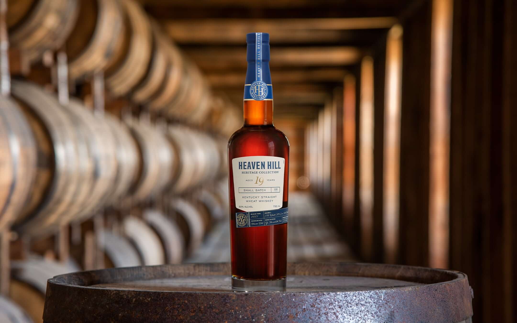 A bottle of Heaven Hill Heritage Collection 19-Year Kentucky Straight Wheat Whiskey displayed on a barrel inside a dimly lit rickhouse, with rows of aging barrels in the background.